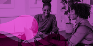 The image shows two black women sitting while having a conversation in front of a laptop.